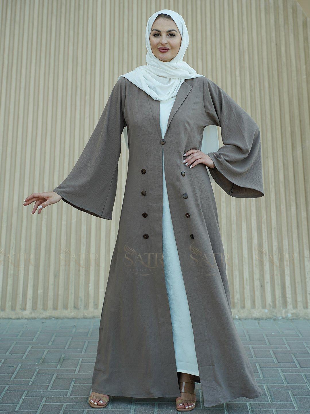 Woman wearing a long, buttoned abaya with a white hijab against a beige wall.