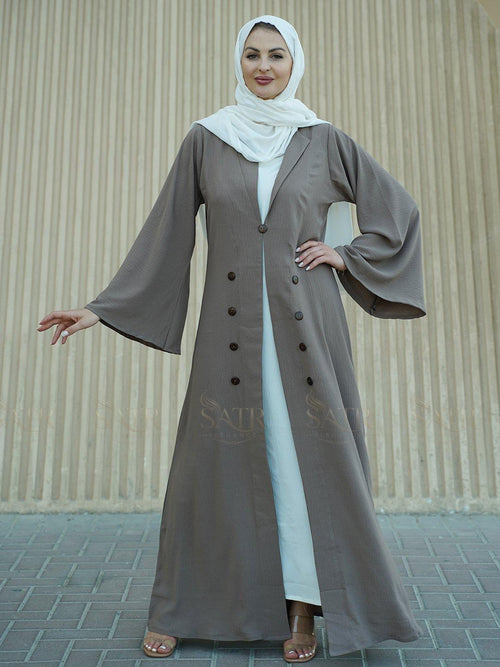 Woman wearing a long, buttoned abaya with a white hijab against a beige wall.