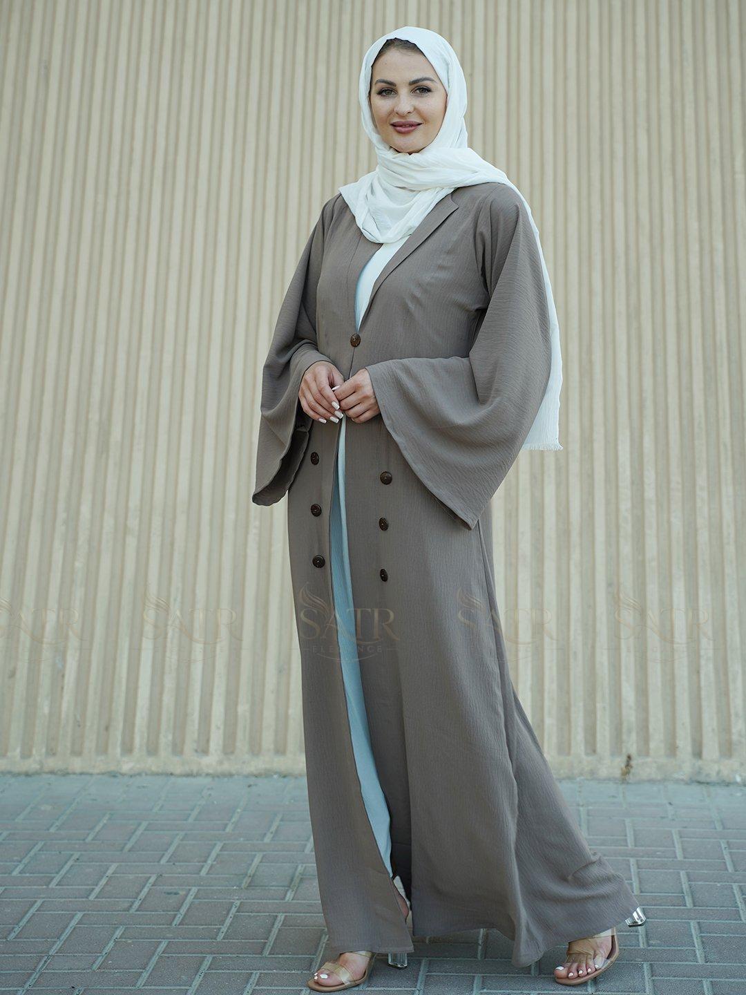Woman wearing a long, light brown dress with a matching hijab against a beige wall.