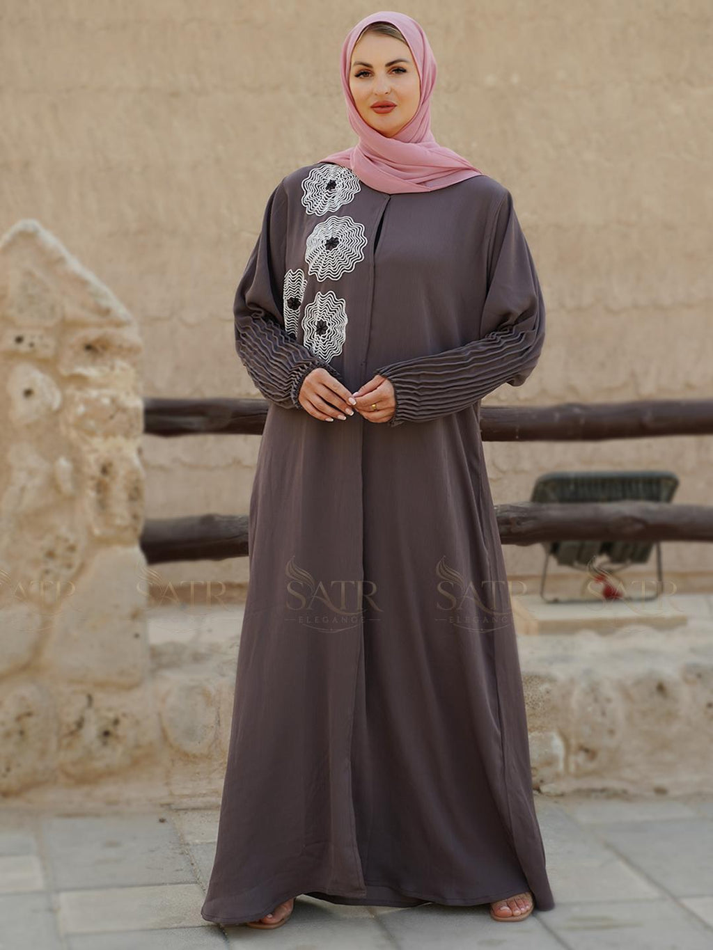 Woman wearing a long brown dress with floral embroidery in front of a stone building.
