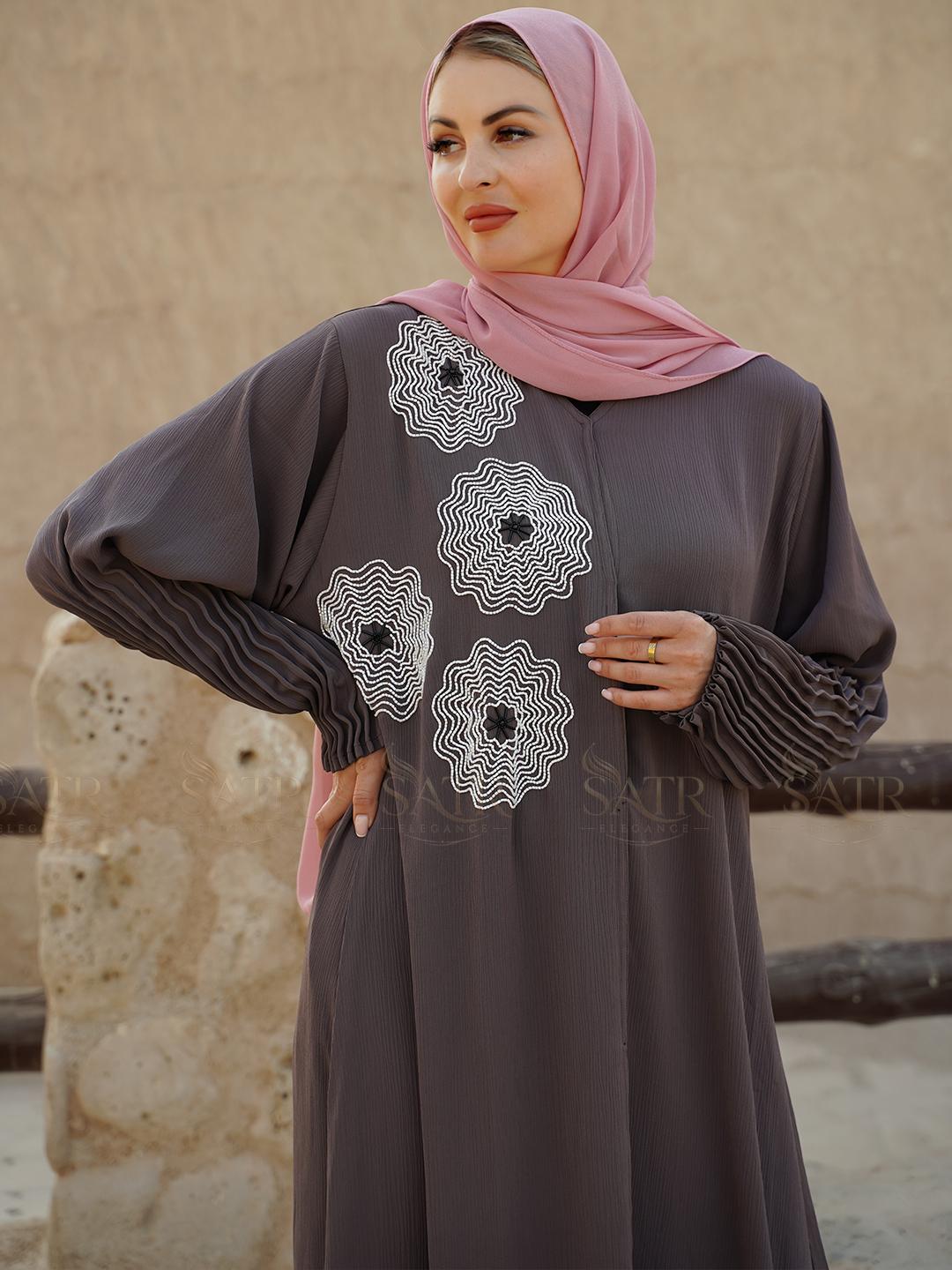 Woman wearing a gray dress with white floral patterns and a pink hijab against a beige wall.