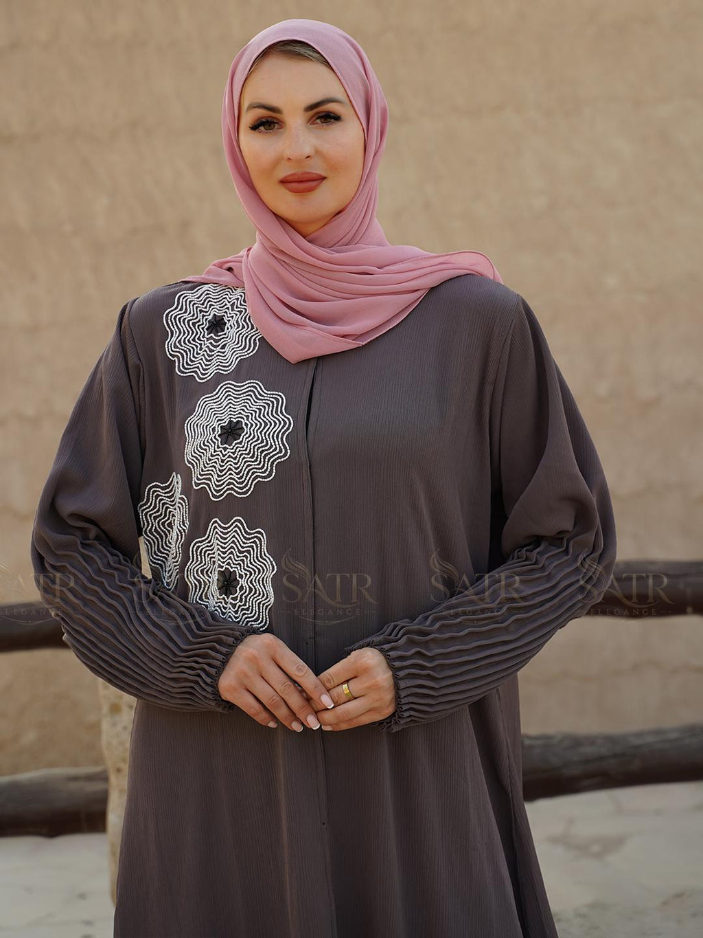 Woman wearing a gray dress with white embroidery and a pink hijab against a beige wall.