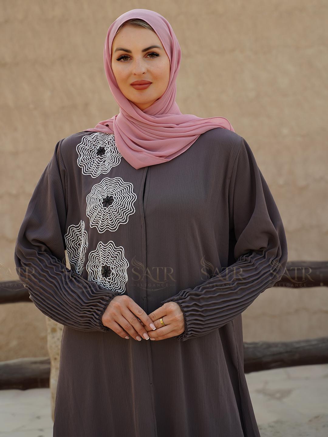 Woman wearing a gray dress with white embroidery and a pink hijab against a beige wall.