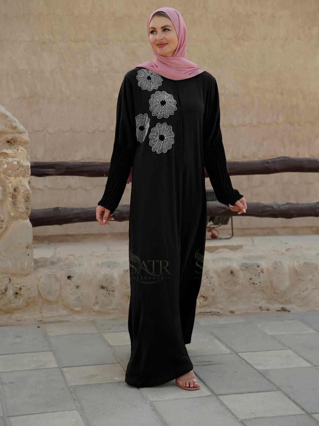 Woman wearing a black dress with floral patterns and a pink hijab standing in front of a stone wall with old dubai vibes