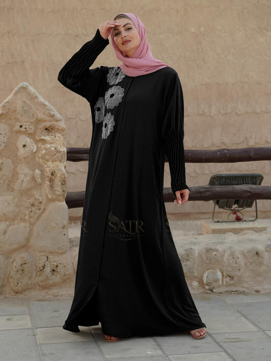 Woman wearing a black abaya with floral patterns in front of a stone wall.
