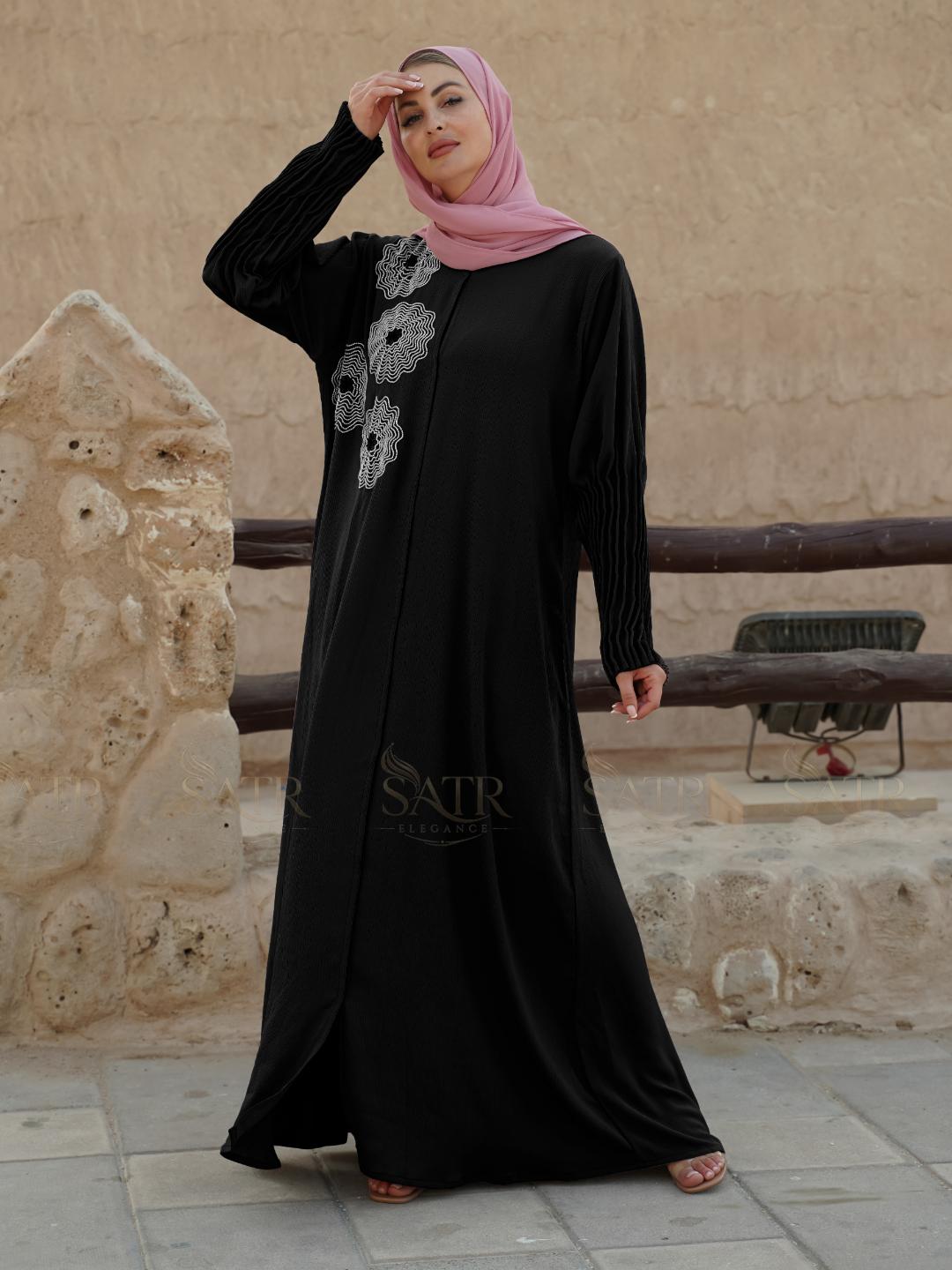 Woman wearing a black abaya with floral patterns in front of a stone wall.