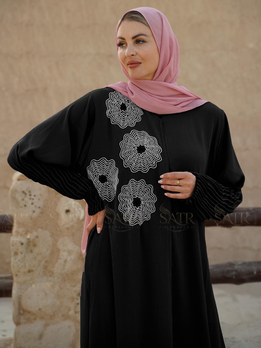 Woman wearing a black dress with white embroidery and a pink hijab against a stone wall background