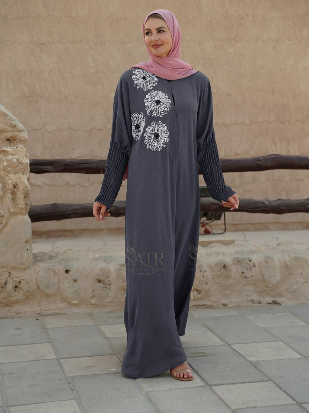 Woman wearing a long dark gray dress with floral patterns in front of a stone wall.