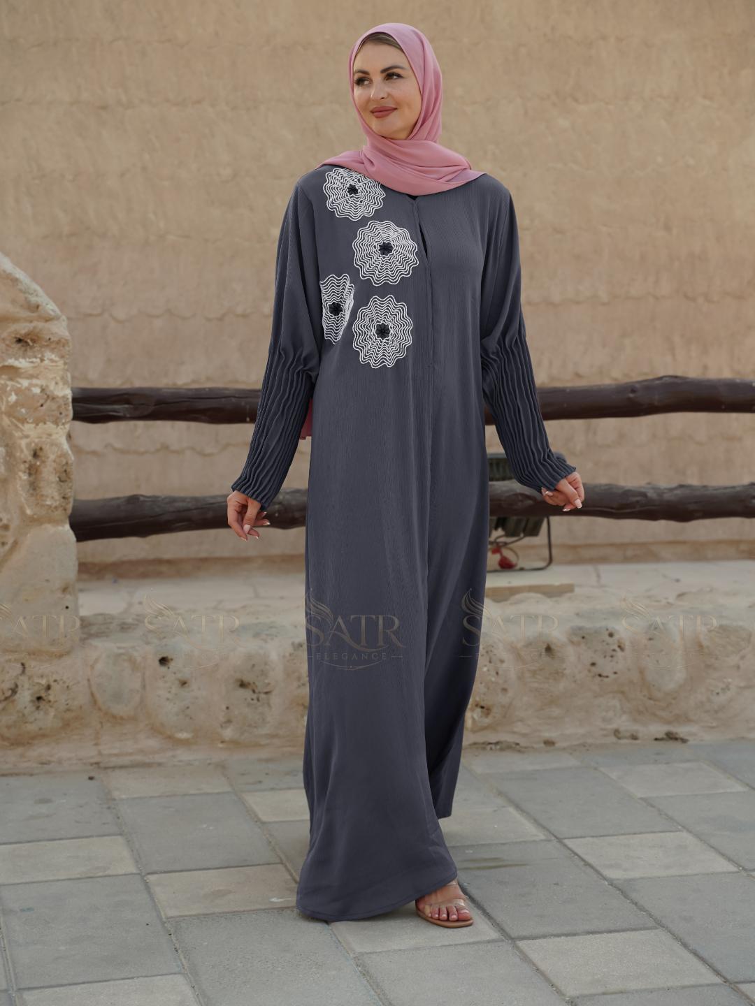 Woman wearing a long dark gray dress with floral patterns in front of a stone wall.