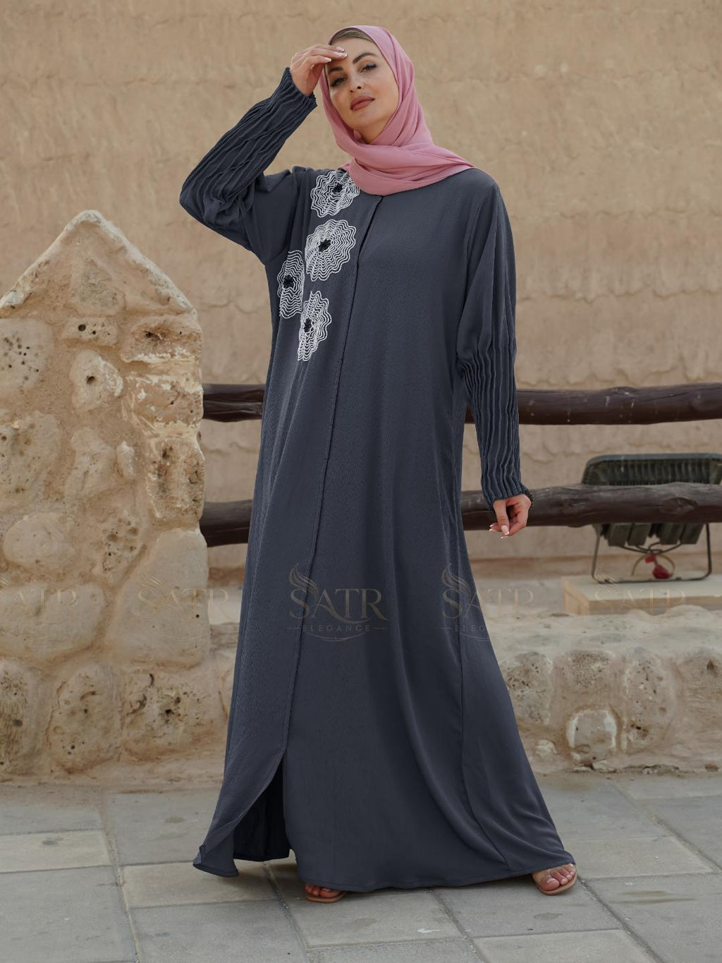 Woman wearing a dark gray abaya with floral patterns in front of a stone wall.