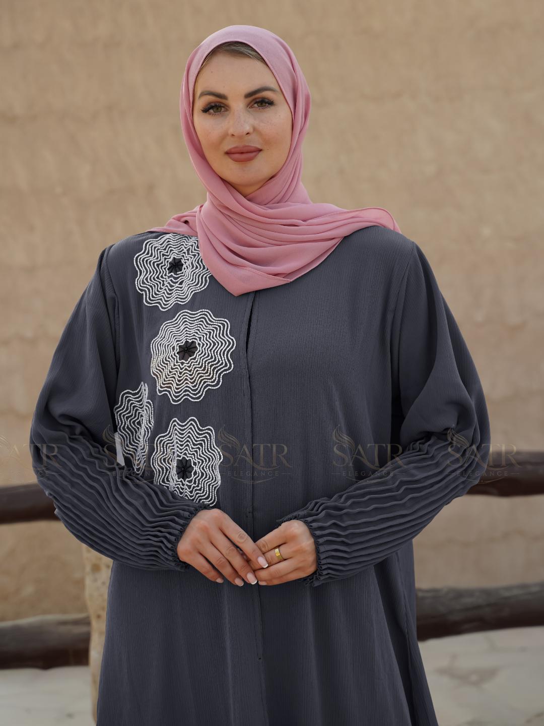 Woman wearing a dark gray dress with white embroidery and a pink hijab against a beige wall.