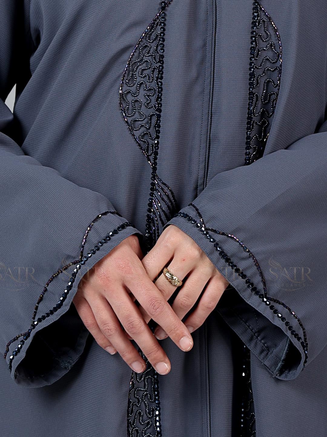 Close-up of a person wearing a grey abaya with intricate black embroidery, focusing on the hands and sleeves.