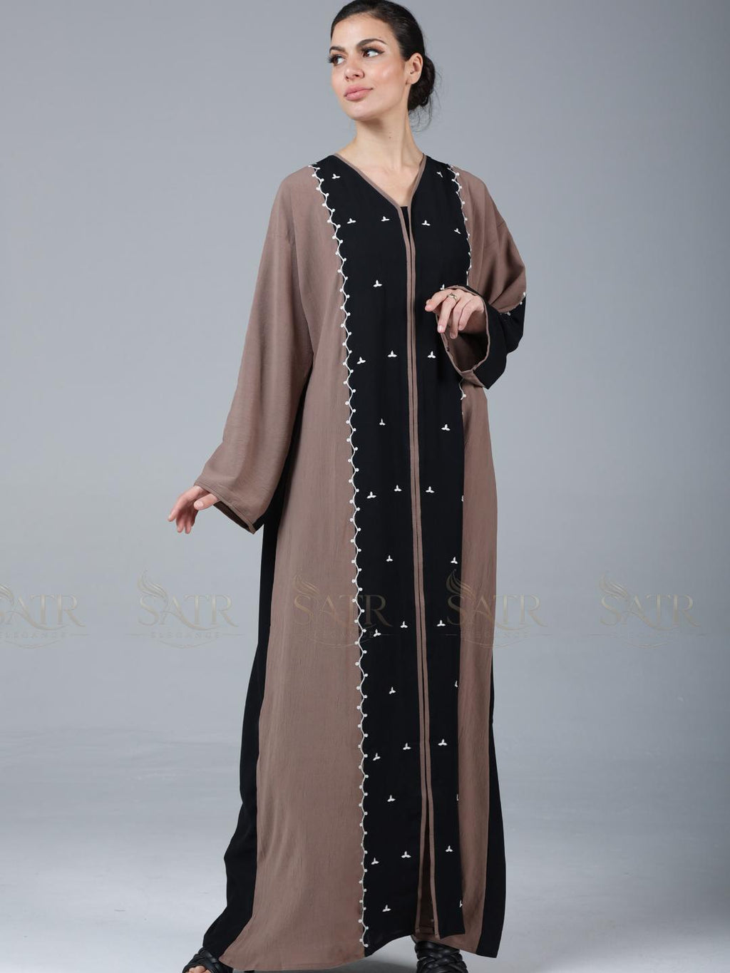 Woman wearing a long, brown and black dress with white embroidery on a gray background