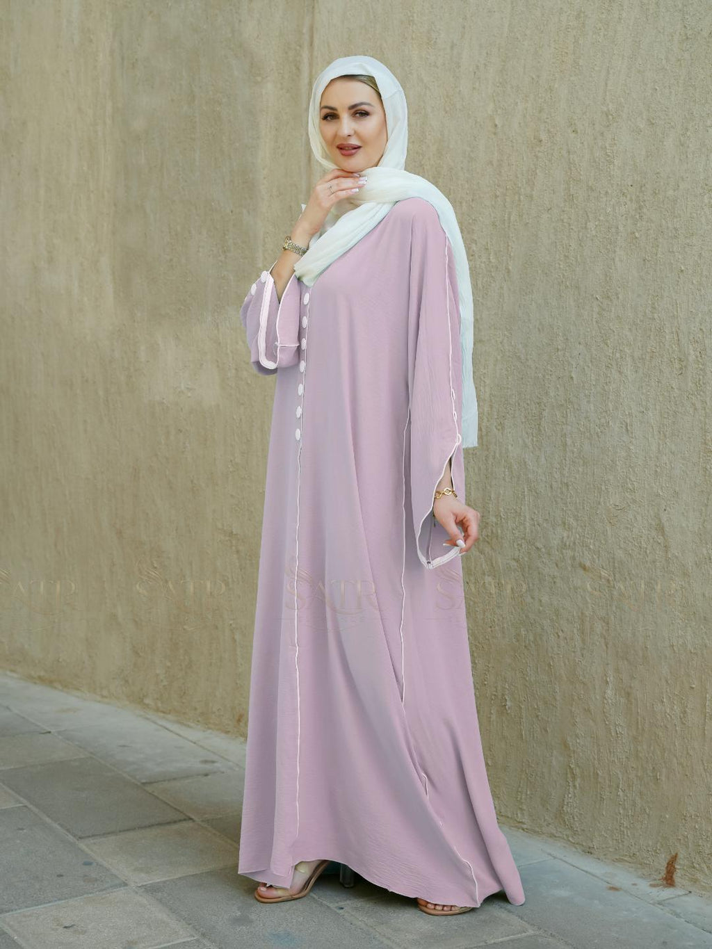 Woman wearing a light pink abaya with a white hijab against a beige wall.