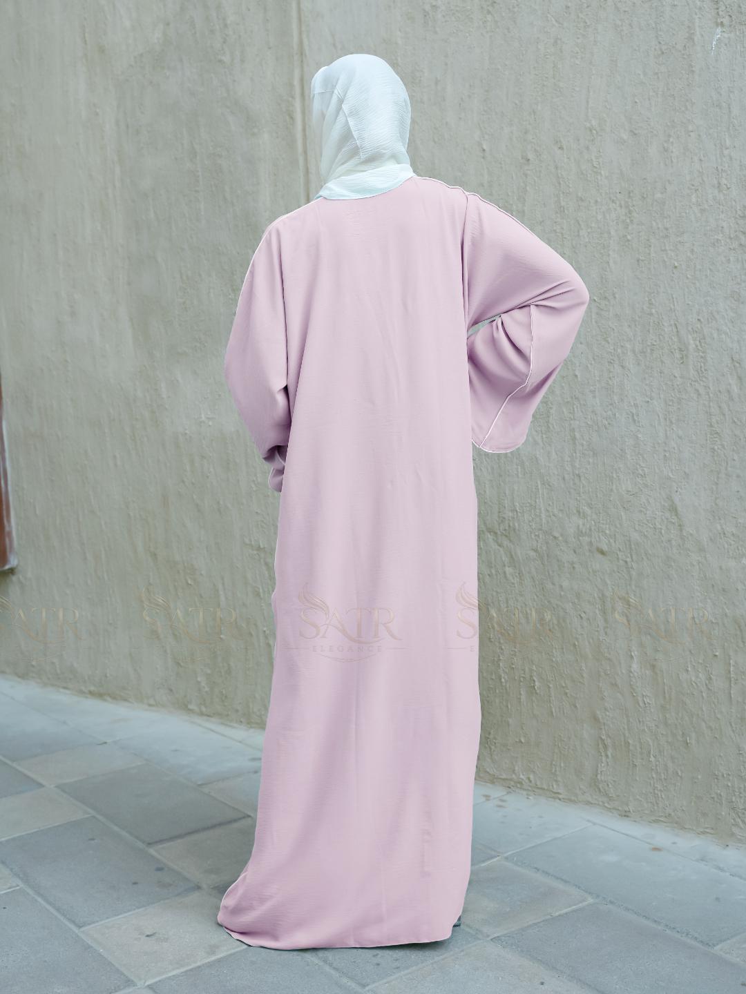 Person wearing a light pink abaya and white hijab against a plain wall.
