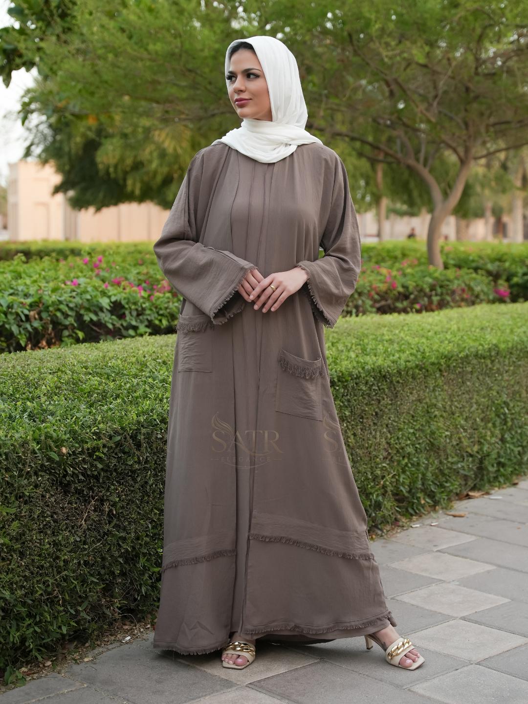 Woman in a brown abaya with a white hijab standing outdoors on a path with greenery.