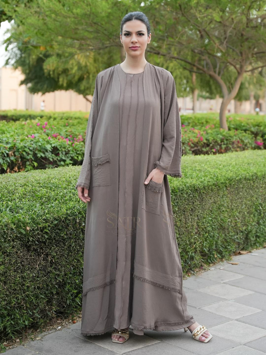 Woman wearing a long, light brown dress standing outdoors with greenery in the background