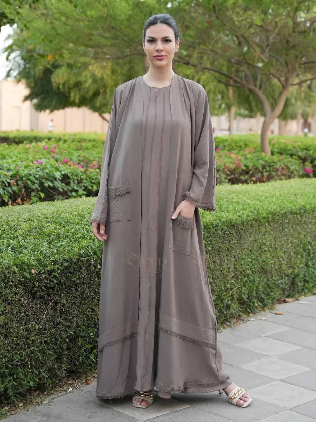 Woman wearing a long, light brown dress standing outdoors with greenery in the background
