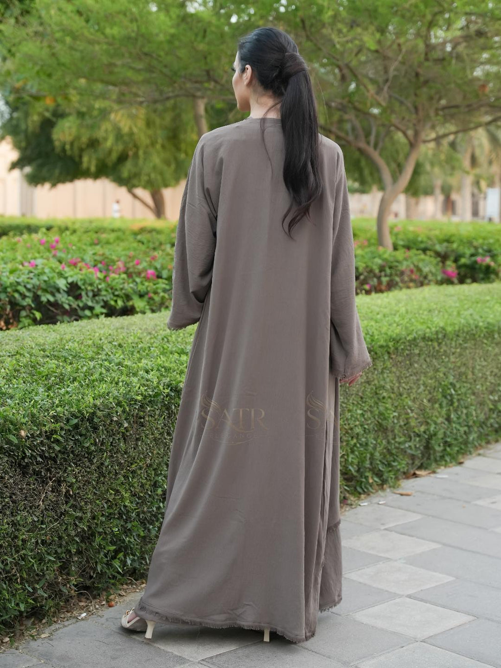 Woman in a long, light brown dress walking on a path with greenery in the background